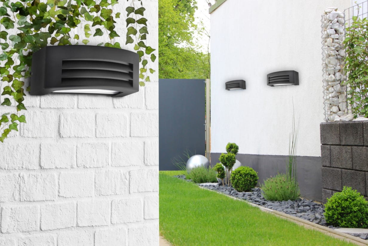 Kinkiet elewacyjny Corso w kolorze antracytowym, z wykrojem w formie żaluzji, zamontowany na białej ścianie. Widoczny w ogrodzie otoczony roślinnością, obok znajduje się kilka innych kinkietów oraz elementy dekoracyjne jak kulista rzeźba.
