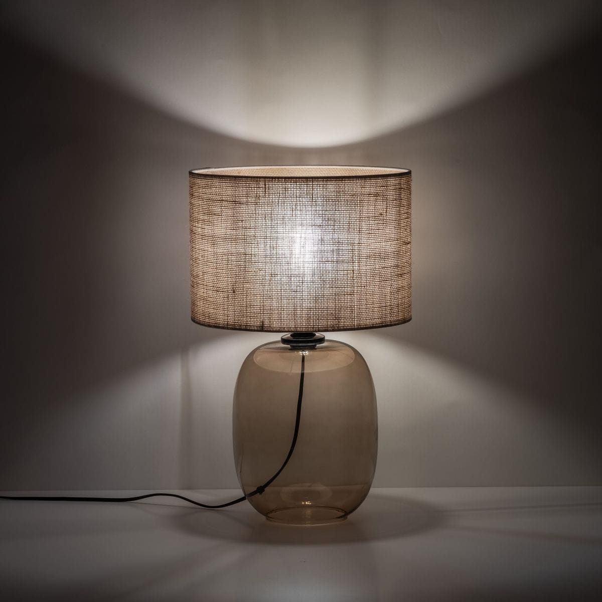 Lampa stołowa Melody z bursztynowym, szklanym korpusem i brązowym, tekstylnym abażurem. Ujęcie frontalne, stojąca, z widocznym przewodem zasilającym. Oświetlenie tworzy ciepłą atmosferę, rzucając delikatne cienie na ścianę.