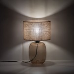 Lampa stołowa Melody z bursztynowym, szklanym korpusem i brązowym, tekstylnym abażurem. Ujęcie frontalne, stojąca, z widocznym przewodem zasilającym. Oświetlenie tworzy ciepłą atmosferę, rzucając delikatne cienie na ścianę.