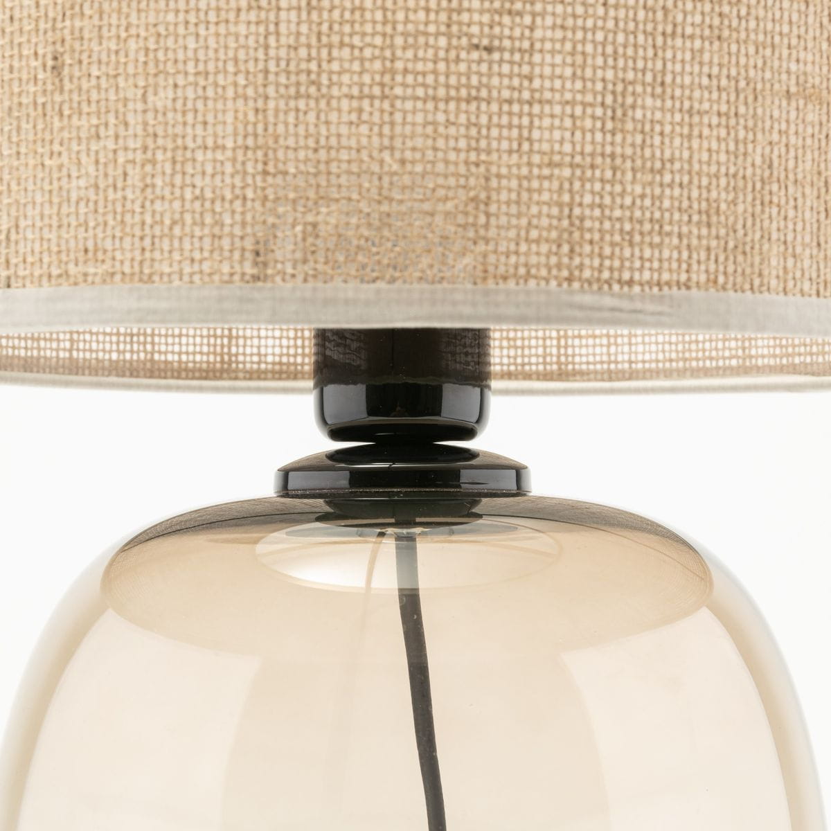 Lampa stołowa Melody z kloszem z jasnego materiału i bursztynową, szklaną podstawą. Widoczny czarny detal u góry. Klosz ma okrągły kształt, a całość prezentuje elegancki styl. Wymiary i detale nie są widoczne.