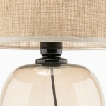 Lampa stołowa Melody z kloszem z jasnego materiału i bursztynową, szklaną podstawą. Widoczny czarny detal u góry. Klosz ma okrągły kształt, a całość prezentuje elegancki styl. Wymiary i detale nie są widoczne.