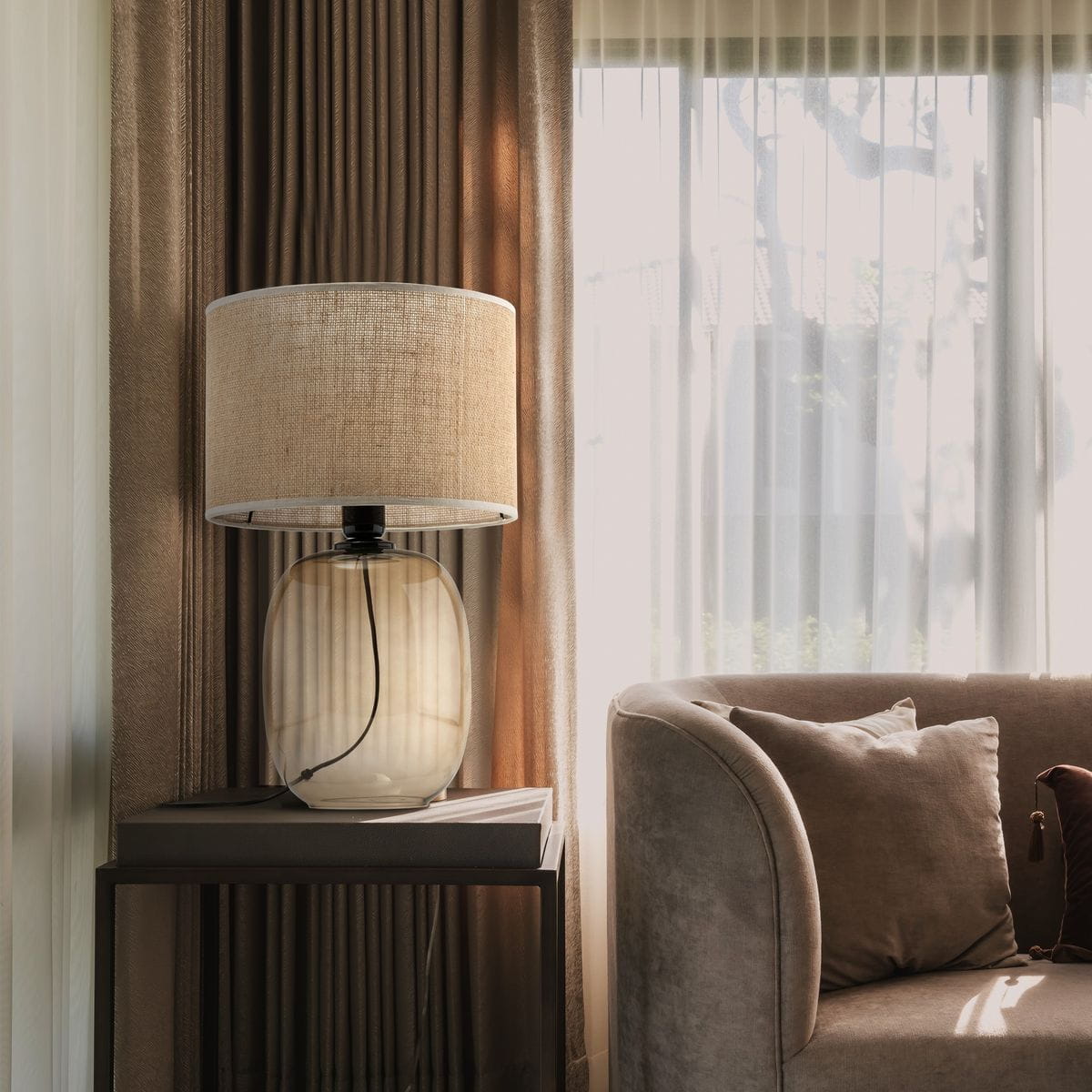 Lampa stołowa Melody z bursztynowym kloszem i brązową podstawą, umieszczona na stoliku obok sofy. Klosz wykonany z naturalnego materiału, z widocznym kablem. Tło z zasłonami w odcieniach brązu, tworzy elegancką aranżację wnętrza.