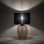 Lampa stołowa Melody z bursztynowym, szklanym kloszem oraz czarnym abażurem. Stoi na płaskiej powierzchni, przewód zasilający widoczny. Klosz tworzy efektowne cienie na ścianie, dodając subtelnego oświetlenia.