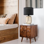 Lampa stołowa Melody z bursztynowym kloszem i czarną oprawą, ustawiona na drewnianym stoliku. Klosz ma okrągły kształt, a lampa znajduje się obok poduszki na łóżku. Tło z elementami drewnianymi i dekoracyjnym obrazem.