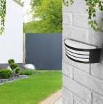 Kinkiet elewacyjny Doris w kolorze czarnym, z białym, prostokątnym kloszem z trzema poziomymi wstawkami. Zamontowany na białej, teksturowanej ścianie, w otoczeniu zieleni i nowoczesnego ogrodzenia. Ujęcie z boku, w naturalnym świetle.