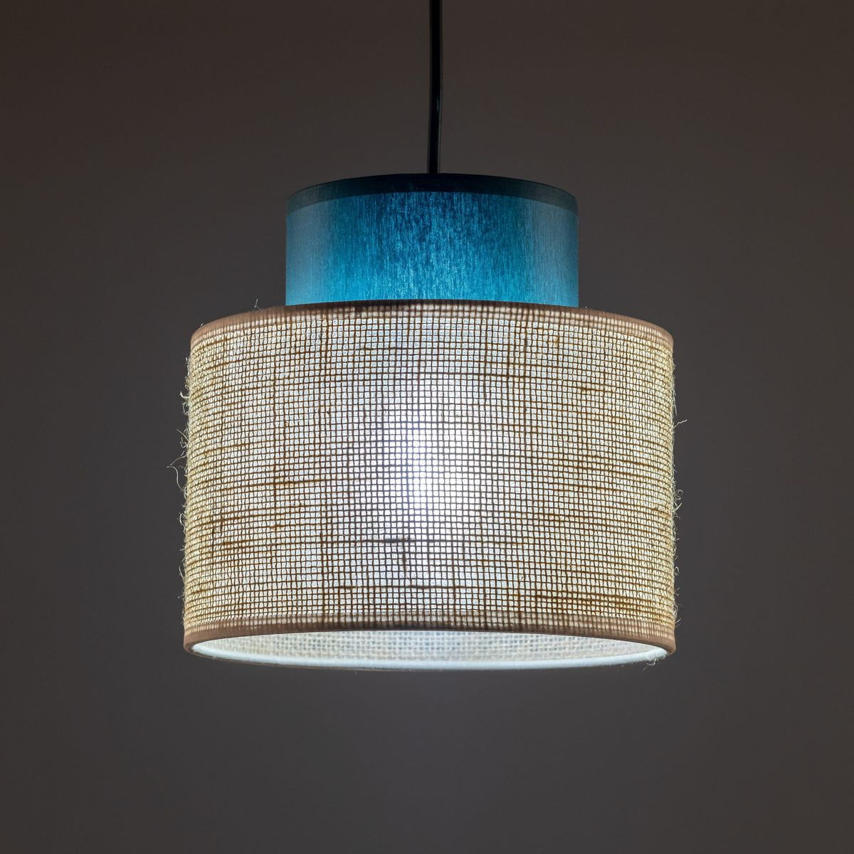 Lampa wisząca Duo D20 z kloszem z juty w naturalnym odcieniu, z niebieską górną częścią. Posiada miejsce na żarówkę E27. Zawieszona pod kątem, w tle ciemne, neutralne kolory.