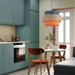 Lampa wisząca Trio 1xE27 w kolorze czarnym z multikolorowymi, okrągłymi kloszami (niebieskim, beżowym i pomarańczowym) zwisa nad okrągłym stołem w nowoczesnej kuchni z zielonymi szafkami i drewnianymi krzesłami.