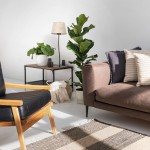 Brązowa lampka stołowa Nicola z abażurem z naturalnego lnu, umieszczona na drewnianym stoliku obok rośliny. W tle widoczna sofa w odcieniu brązowym i czarne krzesło. Jasne tło podkreśla nowoczesny styl aranżacji.