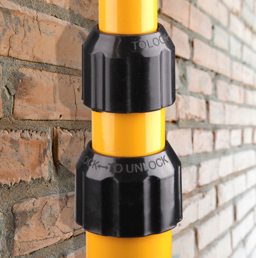 Czarna, plastikowa blokada na żółtym statywie naświetlacza LED. Elementy blokujące są wyraźnie oznaczone napisem „LOCK” i „TO UNLOCK”. Tło stanowi ceglana ściana, co podkreśla konstrukcję produktu.