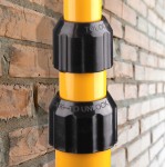 Czarna, plastikowa blokada na żółtym statywie naświetlacza LED. Elementy blokujące są wyraźnie oznaczone napisem „LOCK” i „TO UNLOCK”. Tło stanowi ceglana ściana, co podkreśla konstrukcję produktu.