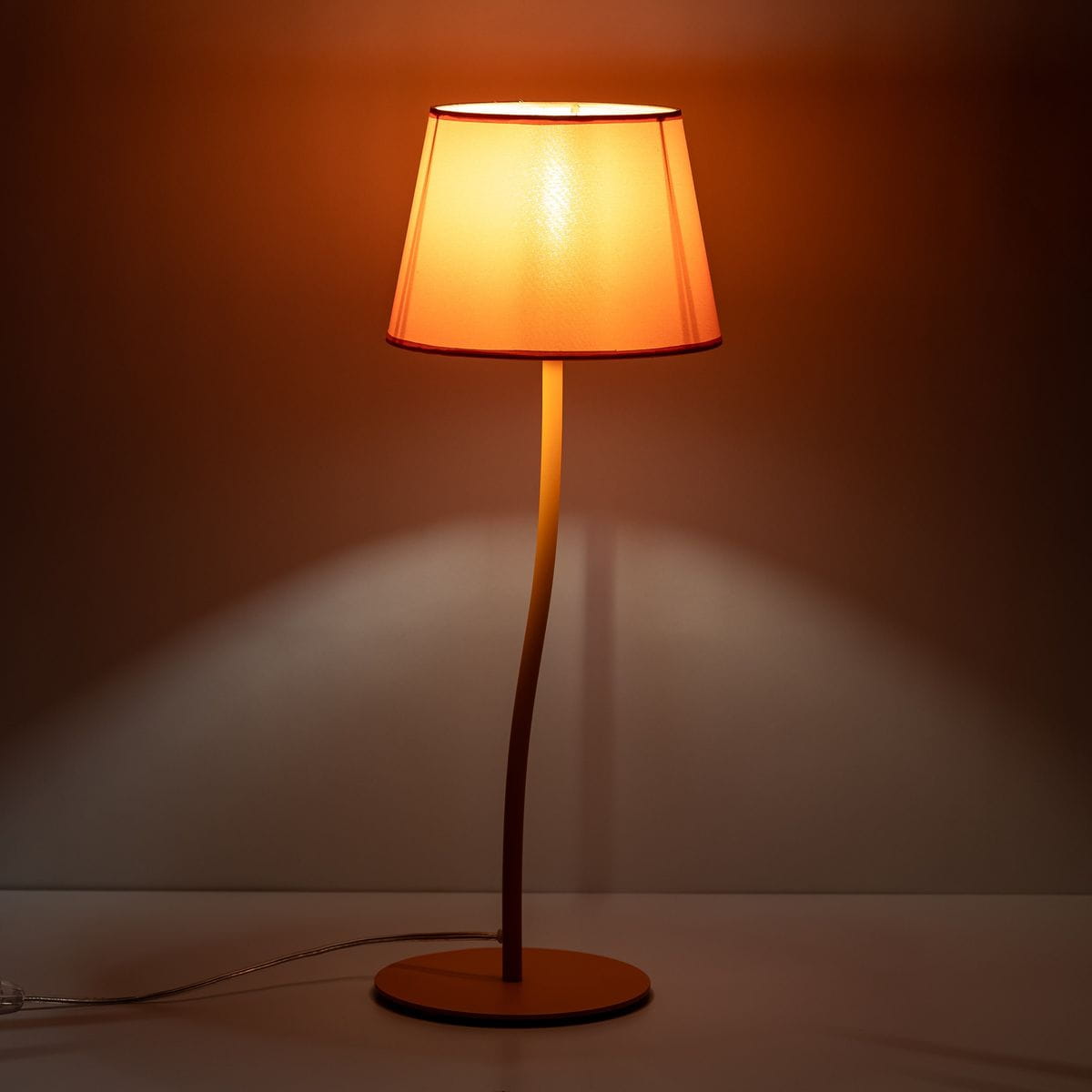 Pomarańczowa lampka stołowa Nicola z kloszem w kształcie stożka, na cienkiej, giętej nodze. Ustawiona na okrągłej podstawie. Ciepłe światło oświetla otoczenie, tworząc przytulną atmosferę. Kabel zasilający widoczny obok.
