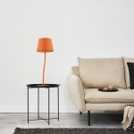 Lampka stołowa Nicola 1xE27 w kolorze pomarańczowym, z metalowym stelażem w czerni i okrągłym abażurem. Ustawiona obok jasnej sofy na czarnym stoliku, w minimalistycznym wnętrzu z białą ścianą.