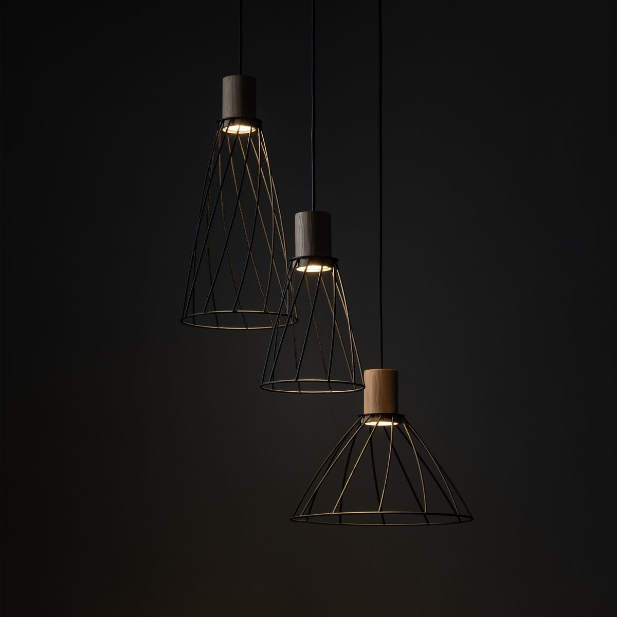 Lampa wisząca Modesto z trzema kloszami w kształcie stożka, wykonanymi z czarnego metalu. Każdy klosz ma drewniane wykończenie u góry. Klosze zawieszone na czarnych przewodach, z delikatnym podświetleniem w ciemnym tle.