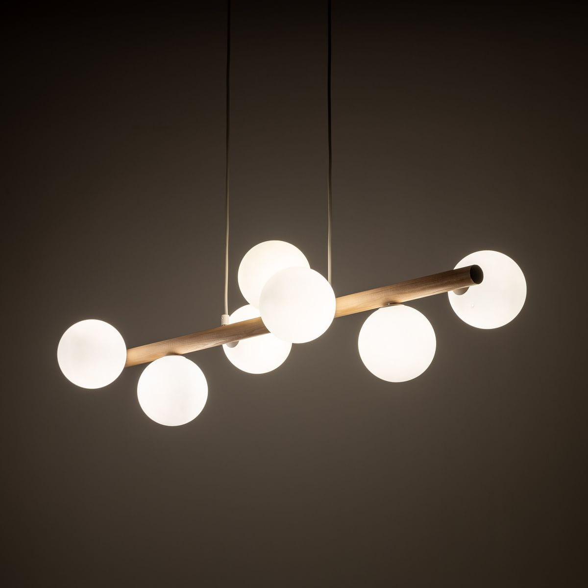 Lampa wisząca Estera z beżowym drewnem, zawieszona poziomo, z siedmioma okrągłymi kloszami z matowego szkła. Klosze rozmieszczone na drewnianej rurze, w jasnym, minimalistycznym stylu, na ciemnym tle.