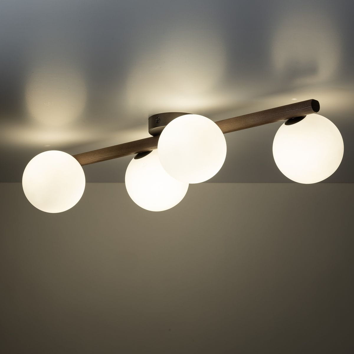Lampa sufitowa Estera z czterema oprawami G9, w brązowym kolorze z ciemnego drewna. Utrzymana w minimalistycznym stylu, z trzema okrągłymi, mlecznymi kloszami, zamontowana na poziomej belce, widok z dołu.