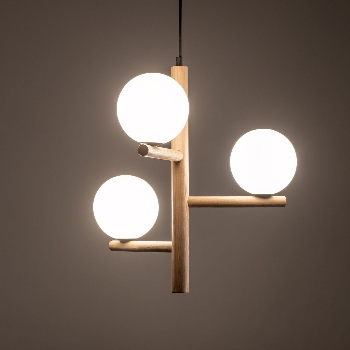 Estera lampa wisząca z trzema okrągłymi kloszami w białym kolorze, zawieszona na beżowym, drewnianym stelażu. Klosze rozmieszczone są asymetrycznie, tworząc nowoczesny wygląd. Lampa zawieszona na cienkim przewodzie na ciemnym tle.