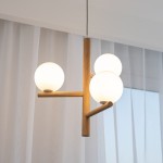 Lampa wisząca Estera z trzema kulistymi kloszami z białego szkła, osadzona na beżowej drewnianej konstrukcji. Klosze umieszczone są na poziomych ramionach, wkomponowane w nowoczesny design.