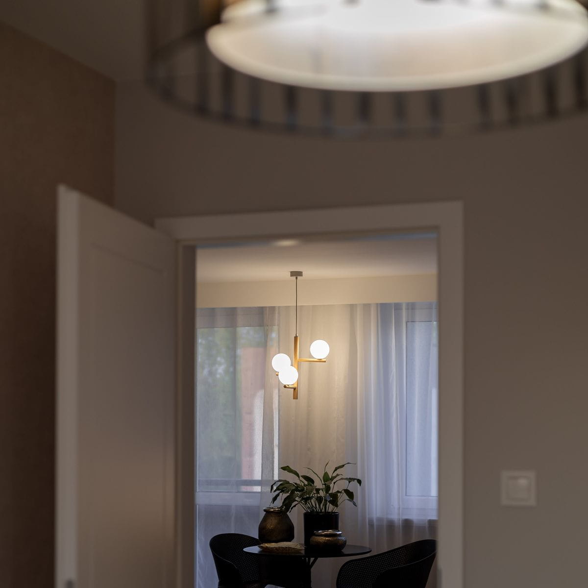 Lampa wisząca Estera w beżowej kolorystyce z jasnym drewnem, z trzema oprawami G9. Produkt zawieszony w pomieszczeniu, z widocznymi kulistymi źródłami światła, na tle okna z firanami i stół z rośliną.