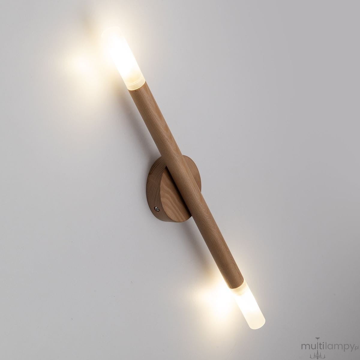 Lampa sufitowa Noble 2xG9 z jasnego drewna, montowana na ścianie. Posiada dwa cylindryczne źródła światła o przezroczystych kloszach, emitujące ciepłe światło. Kąt ujęcia z boku, na jednolitym tle.