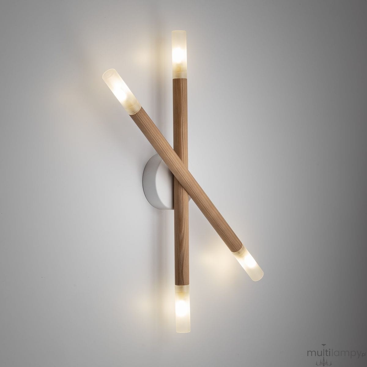 Lampa sufitowa Noble 4xG9 w jasnym drewnie, z dwoma krzyżującymi się elementami. Każdy z nich zakończony jest przezroczystym kloszem, emitującym ciepłe światło. Montaż na ścianie, na tle jasnej powierzchni.
