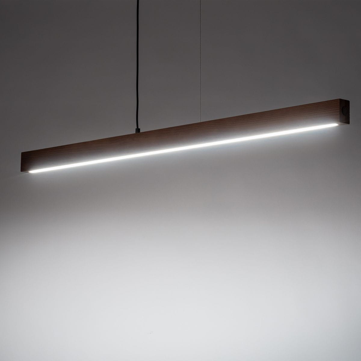 Lampa wisząca Teo o długości prostokątnej, wykonana z ciemnego drewna z czarnym przewodem. Posiada wbudowane oświetlenie LED o mocy 13W, emitujące jasne światło na całej długości. Ujęcie frontalne, zawieszona na dwóch linkach.