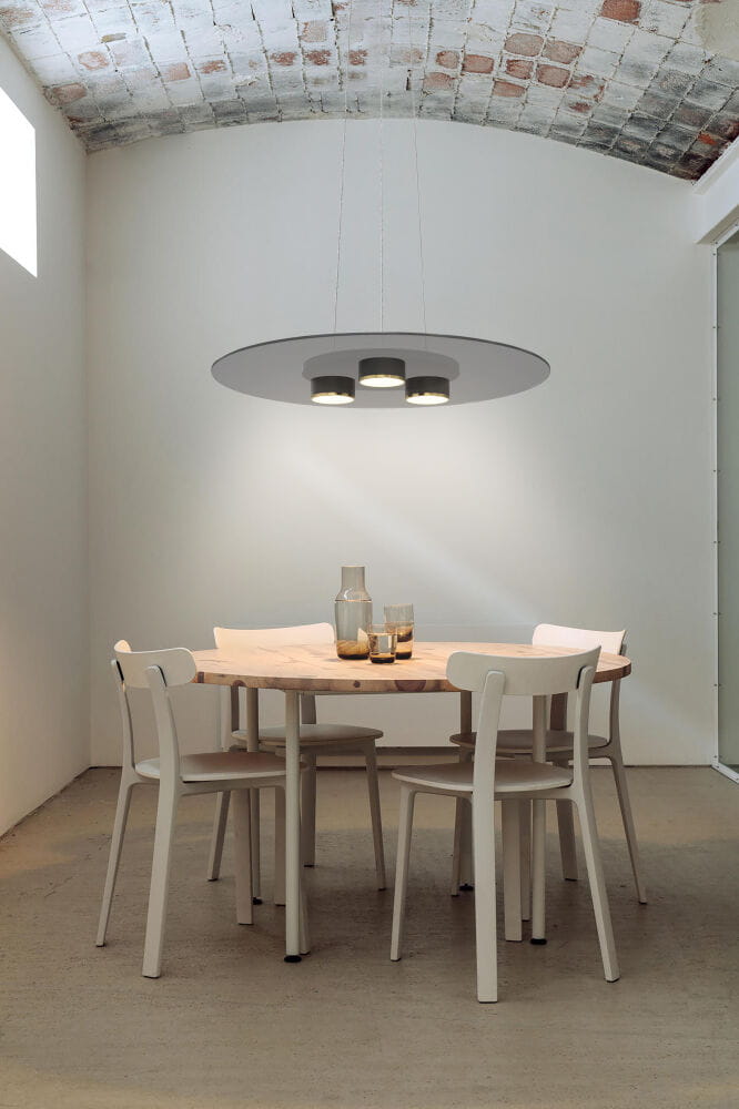 Lannion lampa wisząca w kolorze czarnym i złotym, z trzema źródłami światła LED o mocy 19W. Umieszczona nad okrągłym drewnianym stołem w jasnym wnętrzu, z widocznymi białymi krzesłami. Kąt ujęcia frontalny.