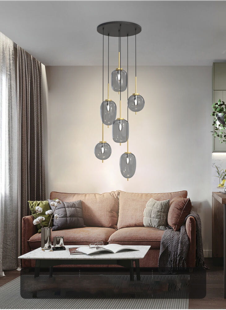 Lampa wisząca Belleville 6xE14 w kolorze czarnym i złotym z sześcioma szklanymi kloszami w odcieniu szarości, zawieszona na długich linkach. Umieszczona w nowoczesnym wnętrzu, nad sofą z poduszkami, w jasnym pomieszczeniu z zasłonami.