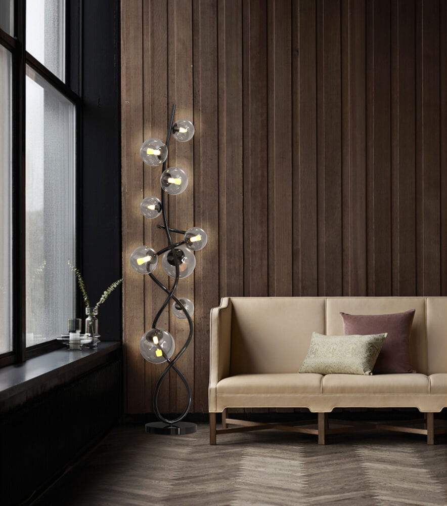 Lampa podłogowa Nancy 9xG9 w kolorze czarnym i chromowanym, z dymionymi kloszami. Ułożona w pozycji stojącej, złożona z wielu ramion. W tle widoczna jasna kanapa z poduszkami w odcieniach szarości i zieleni, umiejscowiona w nowoczesnym wnętrzu.