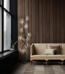 Lampa podłogowa Nancy 9xG9 w kolorze czarnym i chromowanym, z dymionymi kloszami. Ułożona w pozycji stojącej, złożona z wielu ramion. W tle widoczna jasna kanapa z poduszkami w odcieniach szarości i zieleni, umiejscowiona w nowoczesnym wnętrzu.