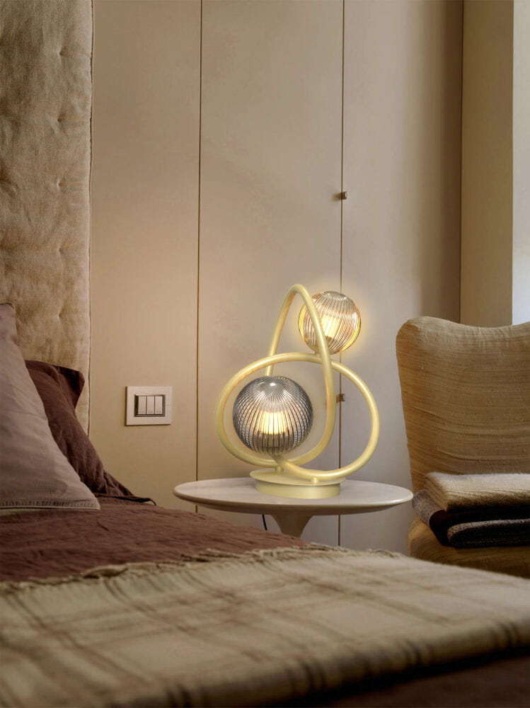 Lampa stołowa Metz 2xG9 w kolorze złotym, z dekoracyjną oprawą w kształcie spirali, umieszczona na białym stoliku obok łóżka. W tle widoczne jasne ściany oraz tekstylia w stonowanych kolorach.