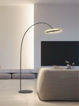 Czarna lampa podłogowa Brest z metalowym, łukowatym ramieniem i okrągłym, złotym kloszem LED. Ustawiona w nowoczesnym wnętrzu obok jasnej sofy, na jednolitym tle. Klosz emituje ciepłe światło, podkreślając elegancki design.