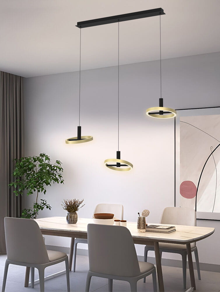 Lampa wisząca Brest z trzema okrągłymi źródłami LED w kolorze czarnym i złotym. Zawieszona na cienkich linkach z prostokątnym, czarnym stelażem. Produkt umieszczony nad drewnianym stołem w nowoczesnym wnętrzu z roślinami i grafiką na ścianie.