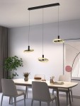 Lampa wisząca Brest z trzema okrągłymi źródłami LED w kolorze czarnym i złotym. Zawieszona na cienkich linkach z prostokątnym, czarnym stelażem. Produkt umieszczony nad drewnianym stołem w nowoczesnym wnętrzu z roślinami i grafiką na ścianie.