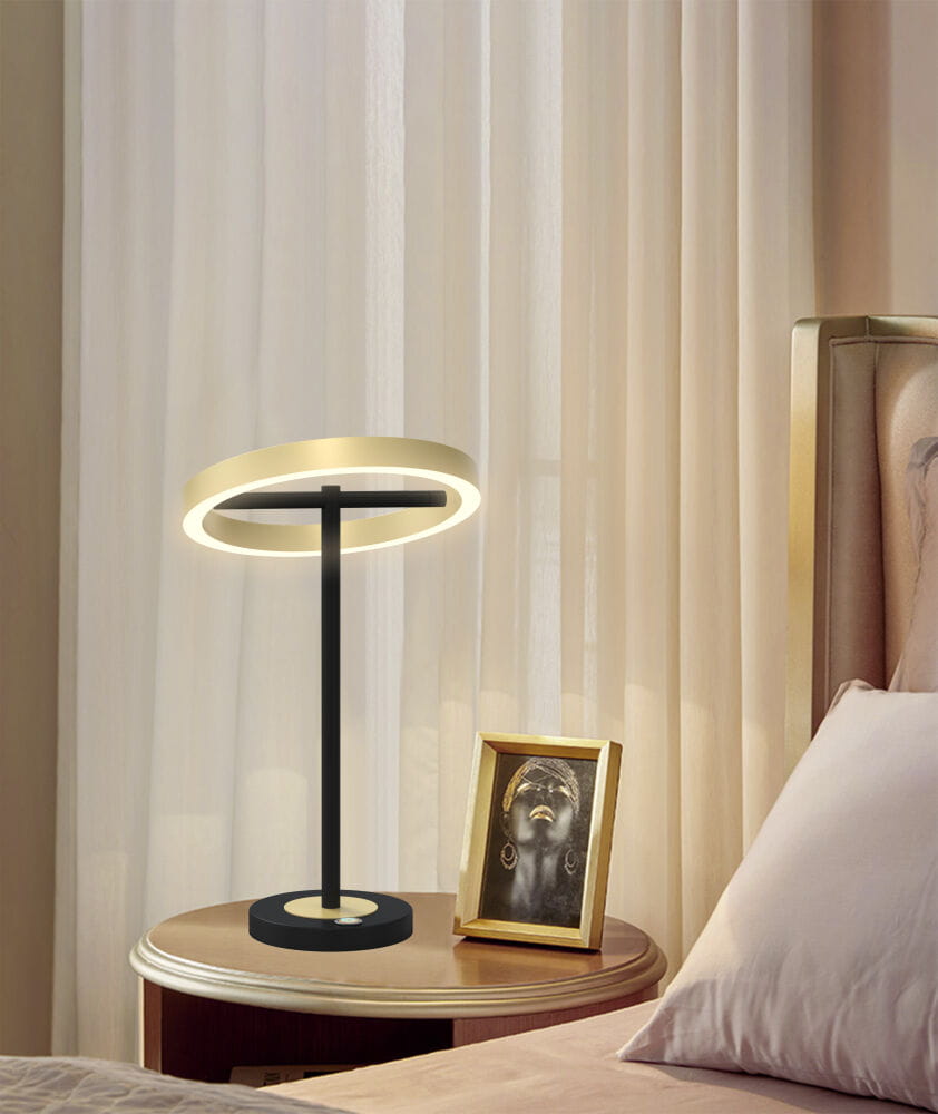 Lampa stołowa Brest z 1xLED/10,5W w kolorze czarnym i złotym. Posiada okrągłą, podświetlaną głowicę na cienkim stelażu, ustawiona na okrągłej podstawie. Umieszczona na stoliku obok ramki ze zdjęciem, w tle zasłony.