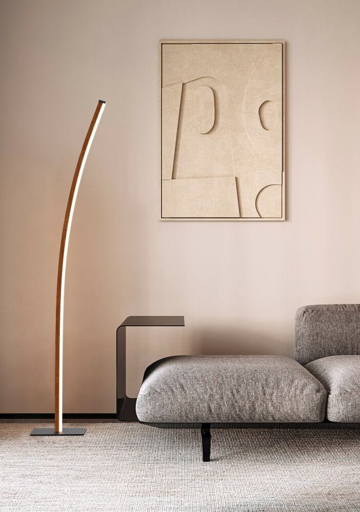 Lampa podłogowa Bologna z czarnym stelażem i drewnianą nogą, o zaokrąglonym kształcie, ustawiona obok szarej sofy i czarnego stolika. Tło w neutralnym kolorze, na ścianie widoczna abstrakcyjna grafika.