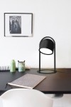 Lampa stołowa Nizza w kolorze czarnym, z okrągłą podstawą i cylindrycznym kloszem, umieszczona na czarnym stole. W tle widoczne akcesoria biurowe oraz czarno-biała fotografia na ścianie. Lampa z jednym źródłem światła LED 10,5W.