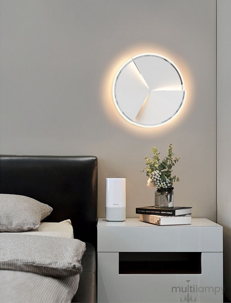 Lampa sufitowa Trapani D49 w chromie z jedną diodą LED 40W, o okrągłym kształcie z trzema białymi panelami, emituje ciepłe światło. Zawieszona na szarej ścianie nad łóżkiem, w nowoczesnym wnętrzu.