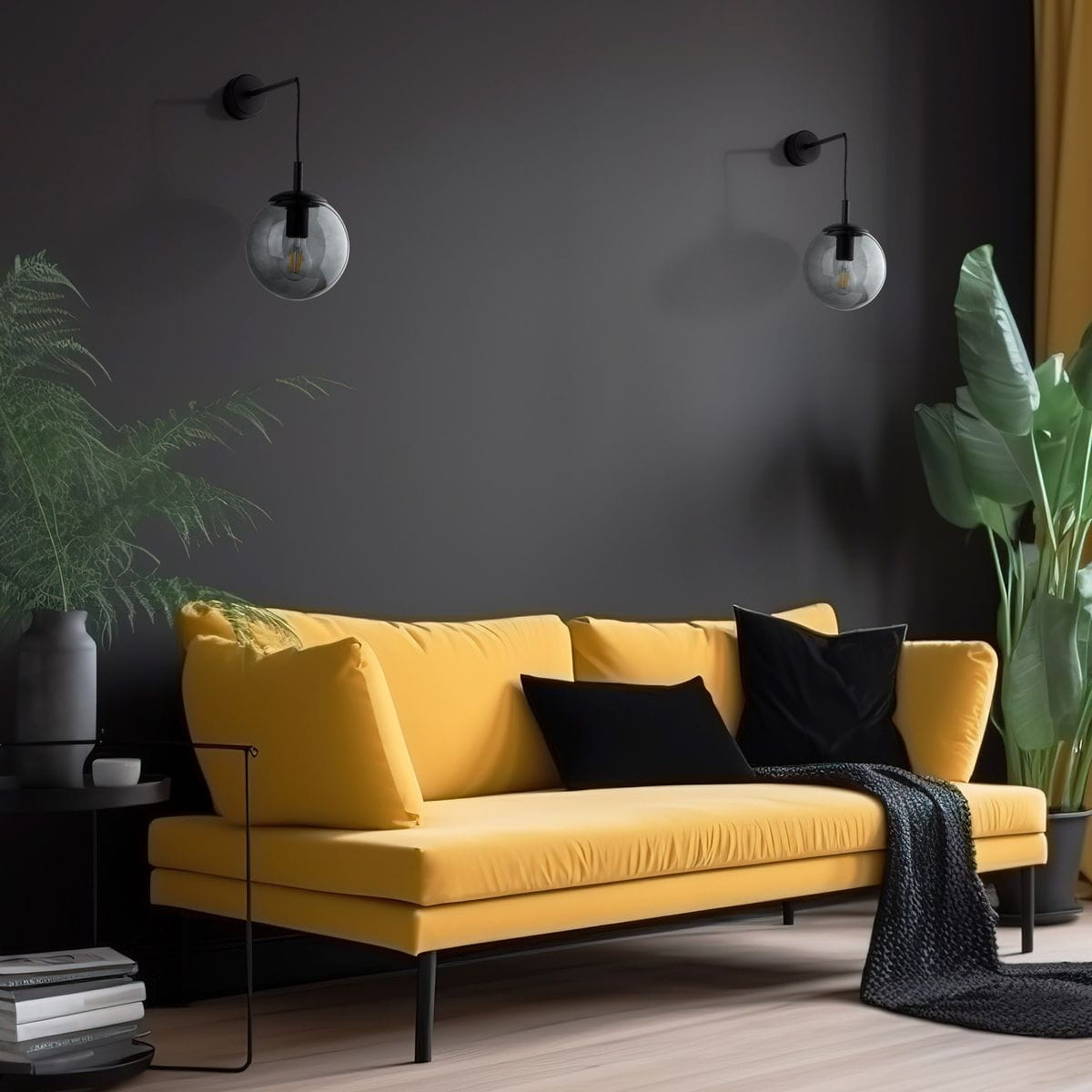 Kinkiet Esme 1xE27 w kolorze czarnym i grafitowym, zamontowany na ciemnej ścianie. Obok żarówki znajduje się przezroczysta kula. W tle żółta sofa z czarnymi poduszkami oraz rośliny dekoracyjne na stoliczku.
