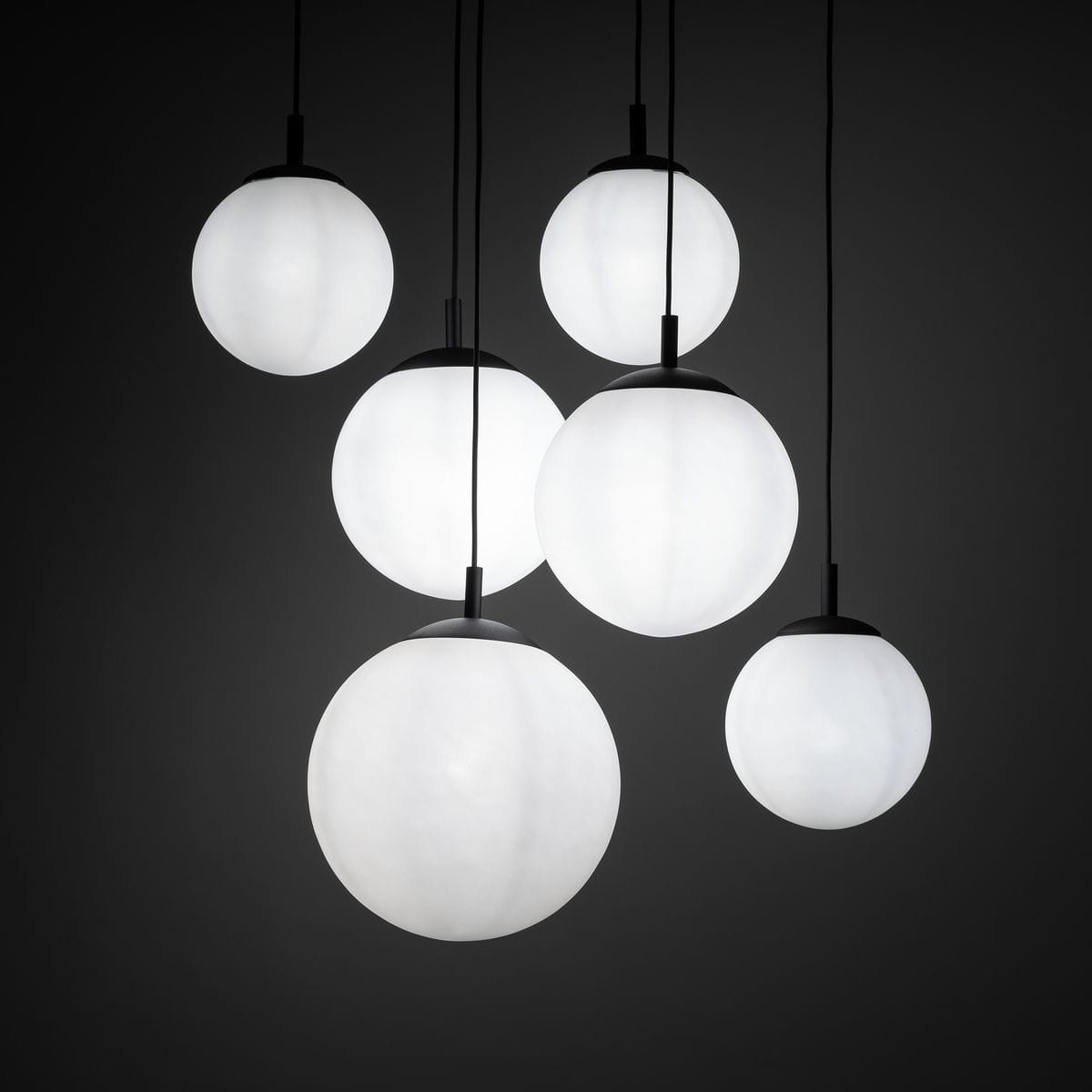 Lampa wisząca Esme z sześcioma okrągłymi kloszami w kolorze białym, zawieszona na cienkich czarnych przewodach. Klosze mają matową powierzchnię i różne rozmiary, tworząc efektowną kompozycję. Tło jest ciemne, co podkreśla jasność lampy.