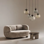 Lampa wisząca Esme z pięcioma kloszami w różnych kolorach, zawieszona na czarnych przewodach. Klosze mają okrągły kształt i są wykonane z przezroczystego szkła. Lampa umieszczona nad jasnoszarym, zaokrąglonym sofą i okrągłym stolikiem.