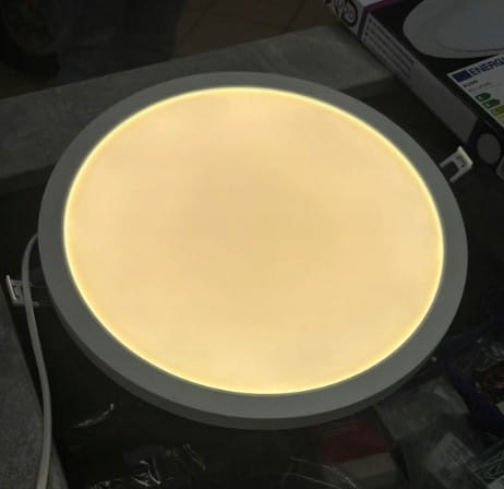 Panel LED D30 8w1/12-24W/3-4-6000K w kształcie okrągłym, natynkowy, biały, z delikatnym podświetleniem. Widok z góry, podłączony do zasilania, umieszczony na szarej powierzchni. Idealny do podtynkowej instalacji oświetleniowej.