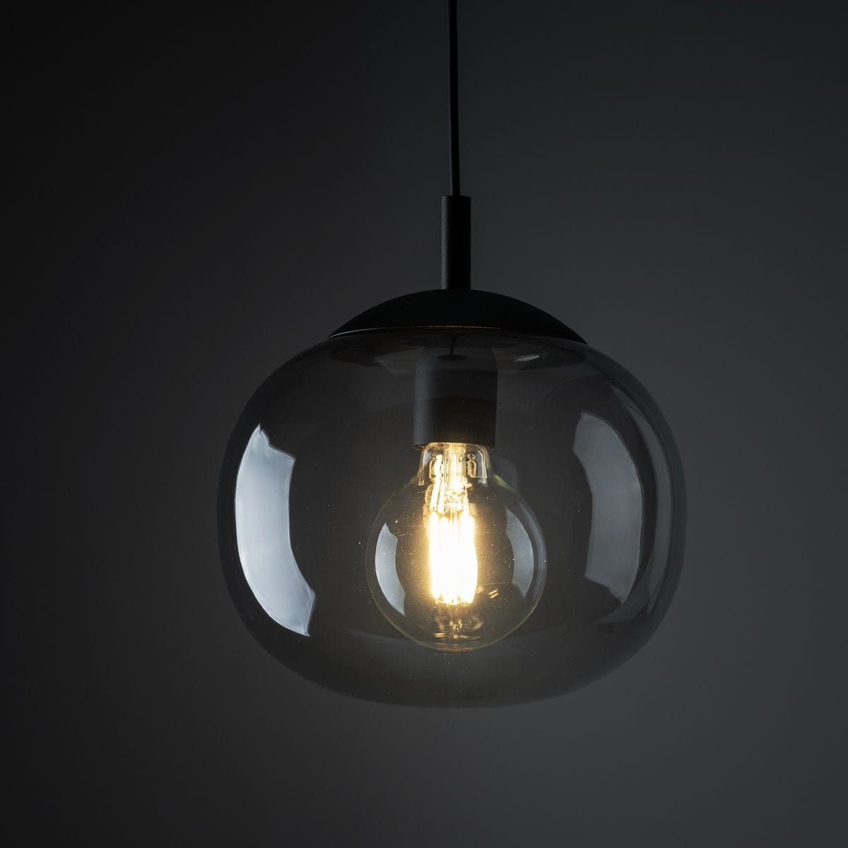 Lampa wisząca Vibe D25 z jedną oprawą E27 w kolorze czarnym i grafitowym, z przezroczystym kloszem, ukazującym żarówkę. Ujęcie z dołu, skupione na detalach konstrukcji i kształcie klosza. Tło ciemne, podkreślające elegancję lampy.