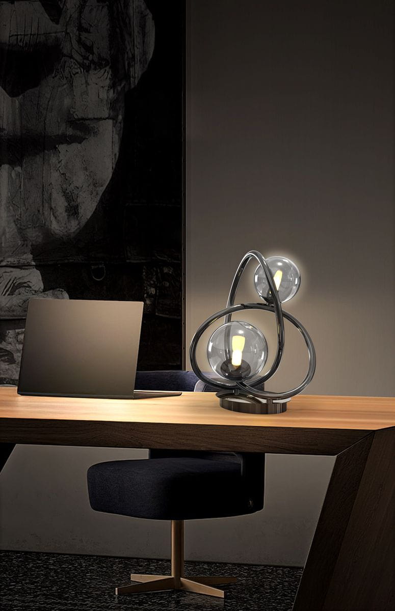 Lampa stołowa Nancy z dwoma źródłami światła G9, w kolorze czarnym z chromowanymi detalami. Okrągłe, dymione klosze umieszczone na eleganckiej podstawie, ustawiona na drewnianym biurku obok laptopa, w minimalistycznym wnętrzu.