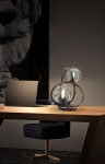 Lampa stołowa Nancy z dwoma źródłami światła G9, w kolorze czarnym z chromowanymi detalami. Okrągłe, dymione klosze umieszczone na eleganckiej podstawie, ustawiona na drewnianym biurku obok laptopa, w minimalistycznym wnętrzu.