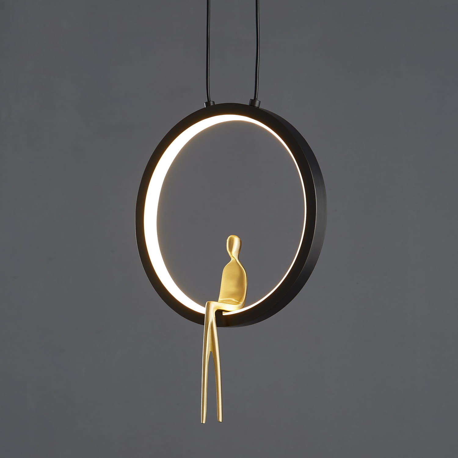 Lampa wisząca Amici z czarnym okrągłym korpusem i mosiężną figurką siedzącą. Źródło światła LED 7W, zawieszona na dwóch cienkich linkach, na szarym tle. Okrągły pierścień podświetlony od wewnątrz, dodający nowoczesnego charakteru.