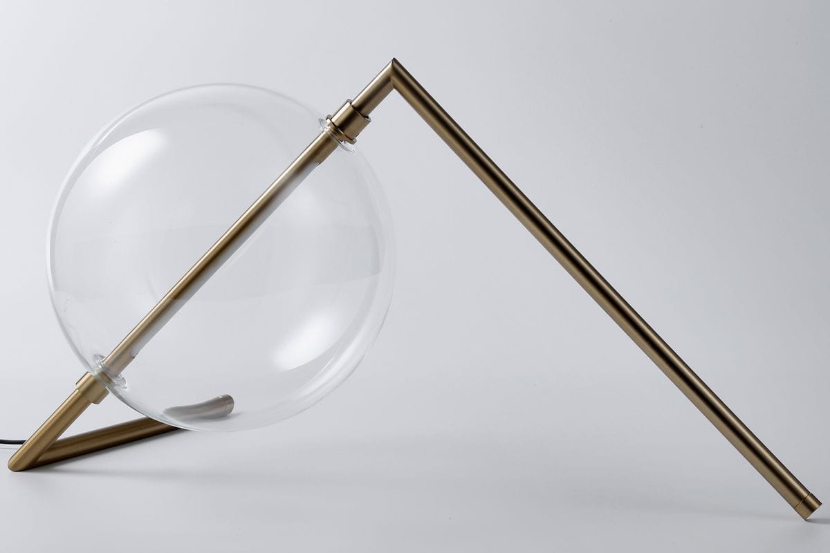 Lampa stołowa Amore z jedną żarówką LED 12W, w złotym wykończeniu. Przezroczysta, okrągła kula w pochyłej pozycji na metalowej podstawie. Kształt minimalistyczny, z widocznym przewodem elektrycznym. Tło jasne, neutralne.