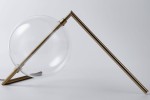 Lampa stołowa Amore z jedną żarówką LED 12W, w złotym wykończeniu. Przezroczysta, okrągła kula w pochyłej pozycji na metalowej podstawie. Kształt minimalistyczny, z widocznym przewodem elektrycznym. Tło jasne, neutralne.
