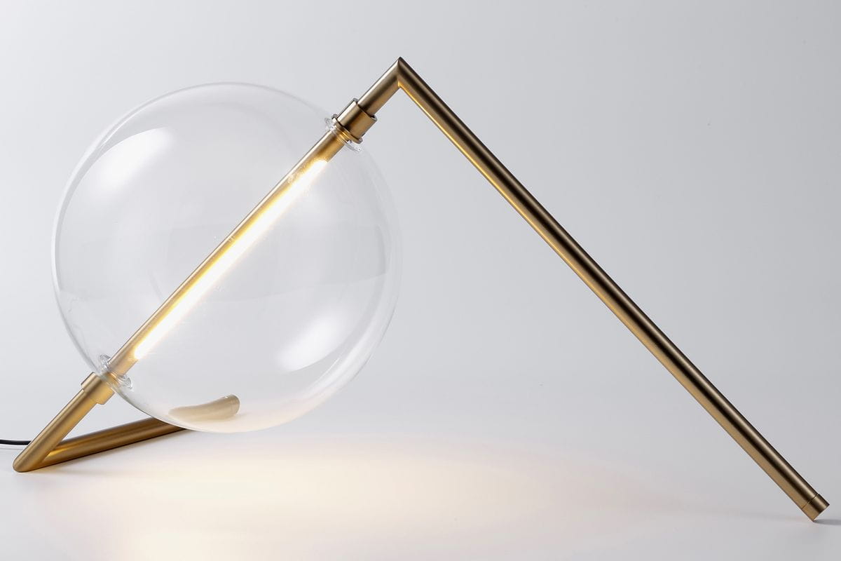 Lampa stołowa Amore z metalowym, złotym stelażem oraz dużą, przezroczystą kulą szklana. Ujęcie z boku, lampa leży na płaskiej powierzchni, z widocznym źródłem światła LED. Minimalistyczny design, idealna do nowoczesnych wnętrz.