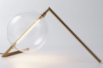 Lampa stołowa Amore z metalowym, złotym stelażem oraz dużą, przezroczystą kulą szklana. Ujęcie z boku, lampa leży na płaskiej powierzchni, z widocznym źródłem światła LED. Minimalistyczny design, idealna do nowoczesnych wnętrz.