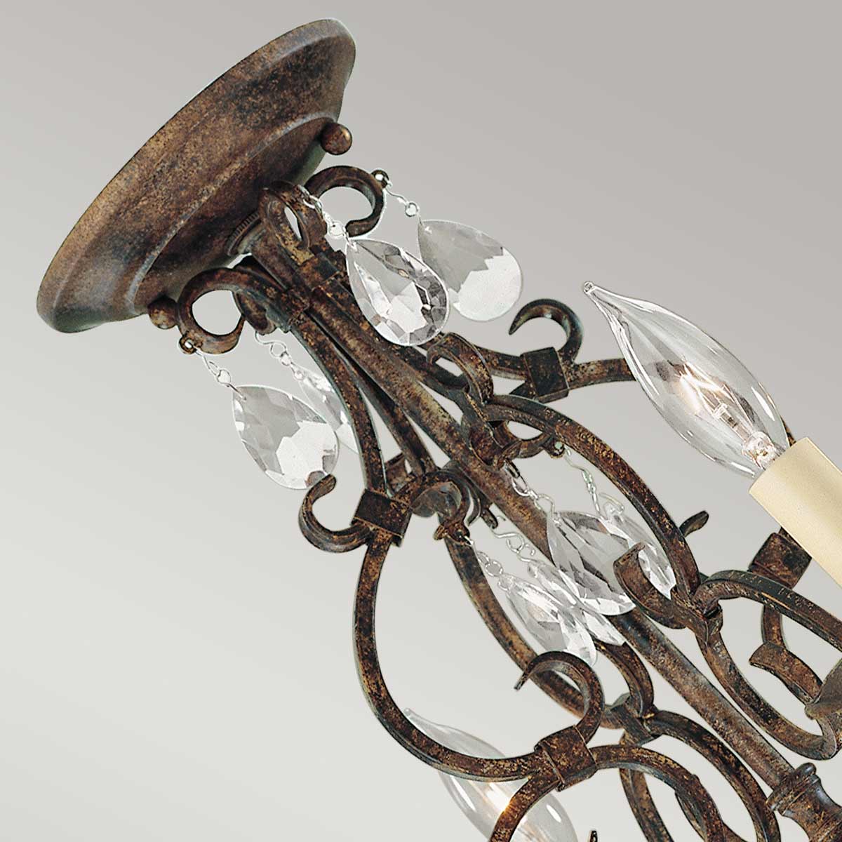 Lampa wisząca Chateau w kolorze brązowym postarzanym, z czterema miejscami na żarówki E14. Ozdobiona przezroczystymi kryształkami w kształcie kropli, z eleganckimi, zakrzywionymi elementami metalowymi. Ujęcie z góry, podkreślające detale konstrukcji.
